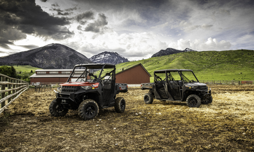 Two Polaris® UTVs parked in the middle of a fenced field next to a barn