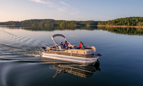 A group of people are boating a SUN TRACKER® pontoon boat on a lake on a sunny day.