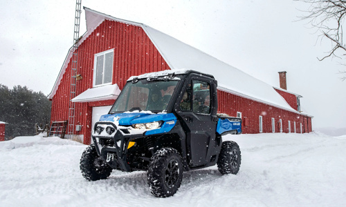 A blue and black Can-Am® Defender® side x side in the snow with a red farmer in the background